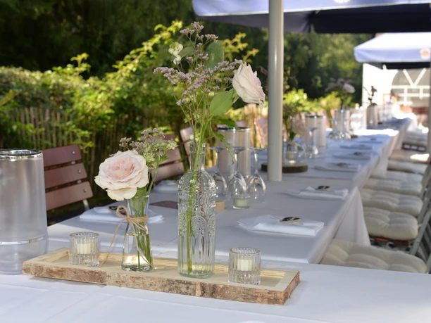 Eine lange, festlich gedeckte Tafel steht unter freiem Himmel, geschmückt mit Blumenarrangements.