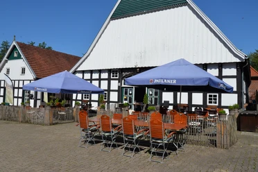 Biergarten auf Nölkenhöner´s Hof Zwei blaue Paulaner-Sonnenschirme beschatten Holztische und -stühle auf einem gepflasterten Hof.