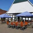 Biergarten auf Nölkenhöner´s Hof Zwei blaue Paulaner-Sonnenschirme beschatten Holztische und -stühle auf einem gepflasterten Hof.