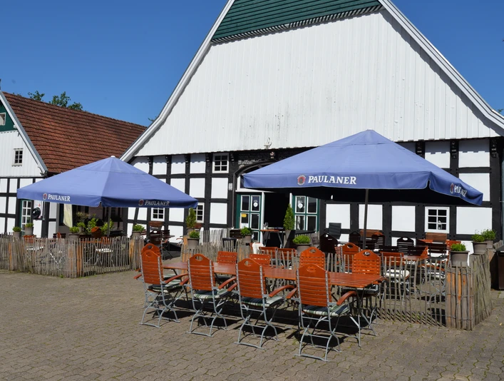 Biergarten auf Nölkenhöner´s Hof Zwei blaue Paulaner-Sonnenschirme beschatten Holztische und -stühle auf einem gepflasterten Hof.