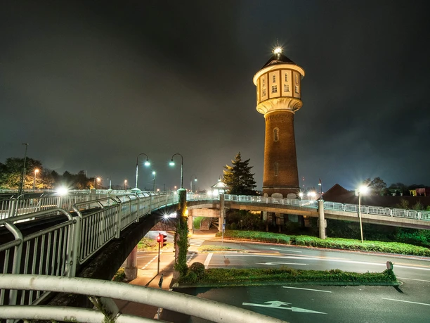 wasserturm-bei-nacht-foto-ludger-h-sken-lh_13110612 Beleuchteter Wasserturm in Lingen bei Nacht, umgeben von Straßen und Fußgängerbrücken.