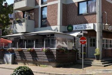 Gasthaus mit rotem Backstein, markantes Jever Deele-Schild über der Eingangstür, Sonnenschirm.