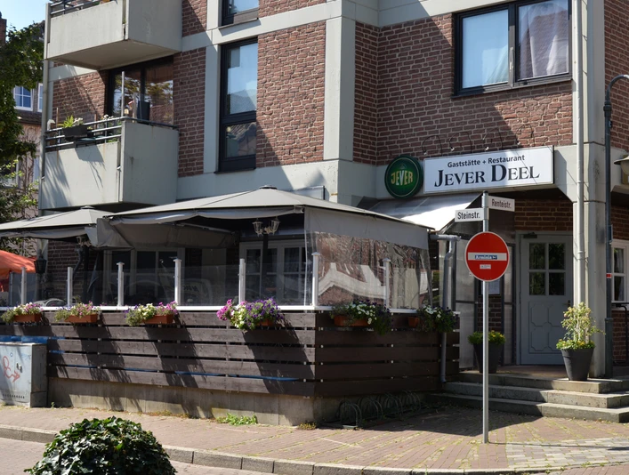 Jever Deele Außenansicht Gasthaus mit rotem Backstein, markantes Jever Deele-Schild über der Eingangstür, Sonnenschirm.
