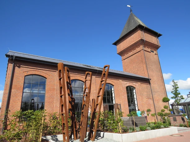 Eisenhütte Backsteinindustriegebäude mit Turm und großen Fenstern vor klarem Himmel; moderne Kunstinstallation vorne.