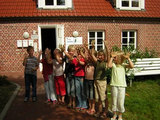 Umweltbildungszentrum Vrees Kindergruppe mit erhobenen Händen steht fröhlich vor einem Backsteingebäude mit rotem Dach.