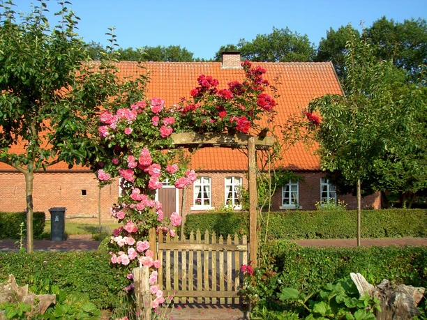 Heimathaus Bauerngarten Oberlangen Backsteinbau mit rotem Ziegeldach, davor blühender Bauerngarten mit Rosenbogen und Holztor