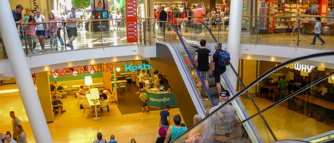Einkaufszentrum mit Rolltreppe, Geschäften, Cafés und vielen Besuchern auf zwei hellen Ebenen