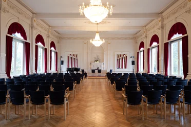 spiegelsaal-bestuhlt Der elegant bestuhlte Spiegelsaal, geprägt vom leuchtenden Kronleuchter und großen Fenstern mit roten Vorhängen.