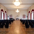 spiegelsaal-bestuhlt Der elegant bestuhlte Spiegelsaal, geprägt vom leuchtenden Kronleuchter und großen Fenstern mit roten Vorhängen.