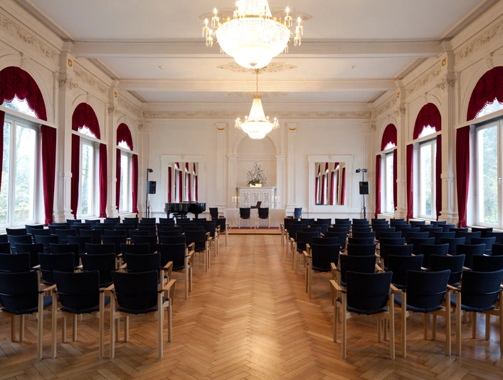 spiegelsaal-bestuhlt Der elegant bestuhlte Spiegelsaal, geprägt vom leuchtenden Kronleuchter und großen Fenstern mit roten Vorhängen.