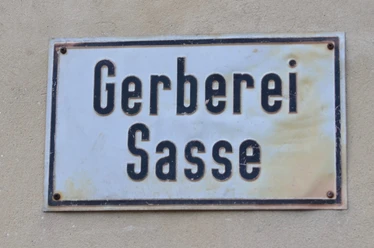 Die alte Gerberei Sasse Historisches Metallschild mit der Aufschrift "Gerberei Sasse" auf beigem Hintergrund.