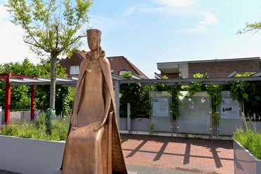 Bronzestatue einer sitzenden Frau in mittelalterlicher Kleidung, umgeben von grüner Bepflanzung.
