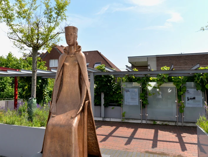 Mathilde-Statur Bronzestatue einer sitzenden Frau in mittelalterlicher Kleidung, umgeben von grüner Bepflanzung.