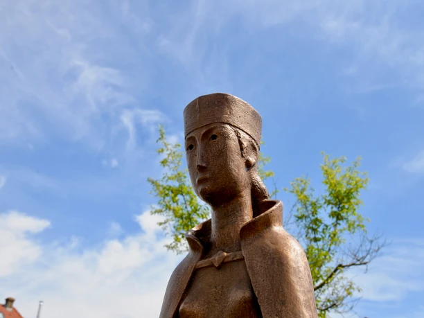 Bronzestatue von Mathilde mit traditionellem Kopfputz, umgeben von blauem Himmel und grünen Blättern.