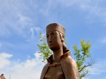 Mathilde Portrait Bronzestatue von Mathilde mit traditionellem Kopfputz, umgeben von blauem Himmel und grünen Blättern.