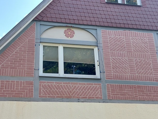 Ein teilrenoviertes Haus mit einem rechteckigen Fenster. Die Fassade ist mit roten und grauen Ziegeln sowie einem schlichten Dach gestaltet.