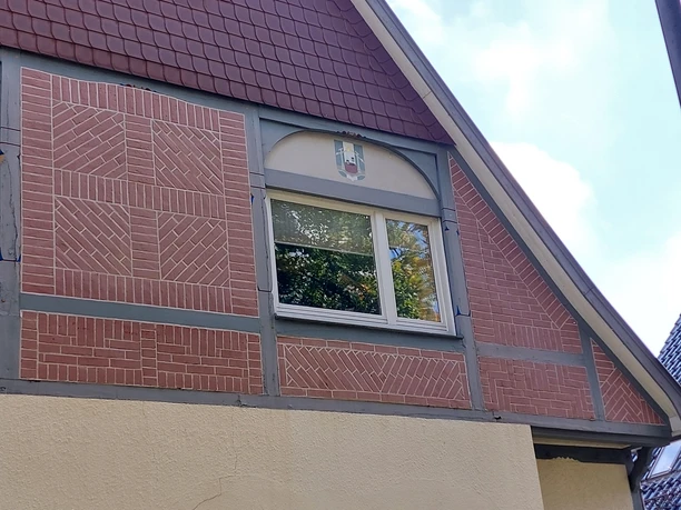 Eine charmante Hausfassade mit dekorativen Mauerwerken und einem Fenster. Der obere Teil des Daches ist in roten Schindeln gedeckt.