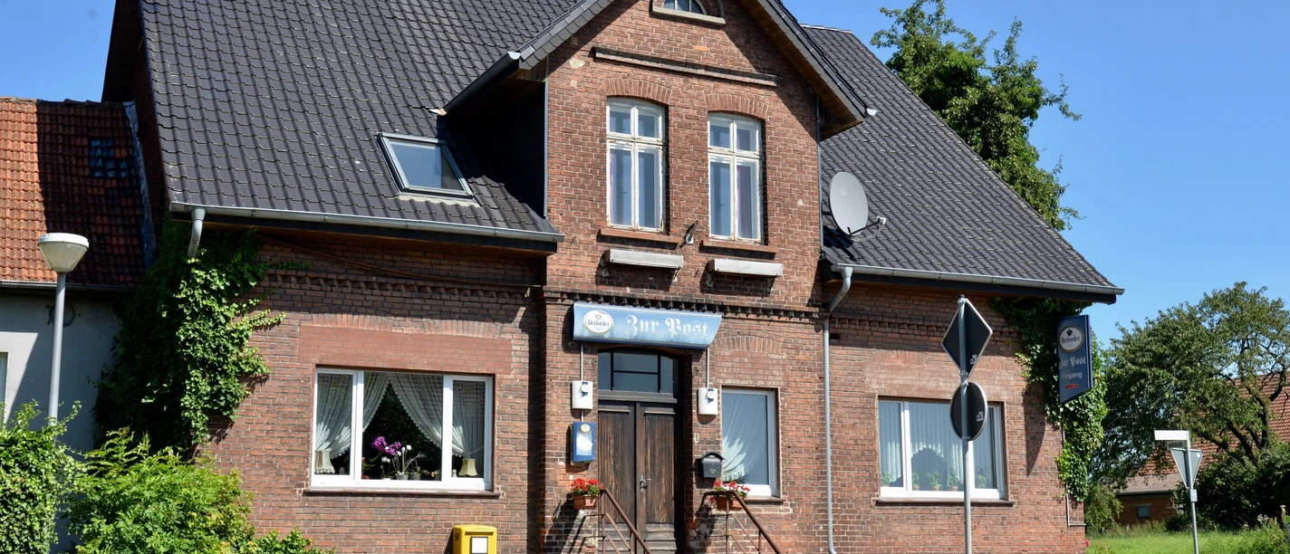 Backstein-Bau mit Ziegeldach und Treppe, zwei Fenster, umgeben von Pflanzen, unter blauem Himmel.