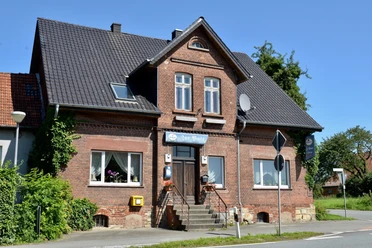 Restaurant Zur Post Backstein-Bau mit Ziegeldach und Treppe, zwei Fenster, umgeben von Pflanzen, unter blauem Himmel.