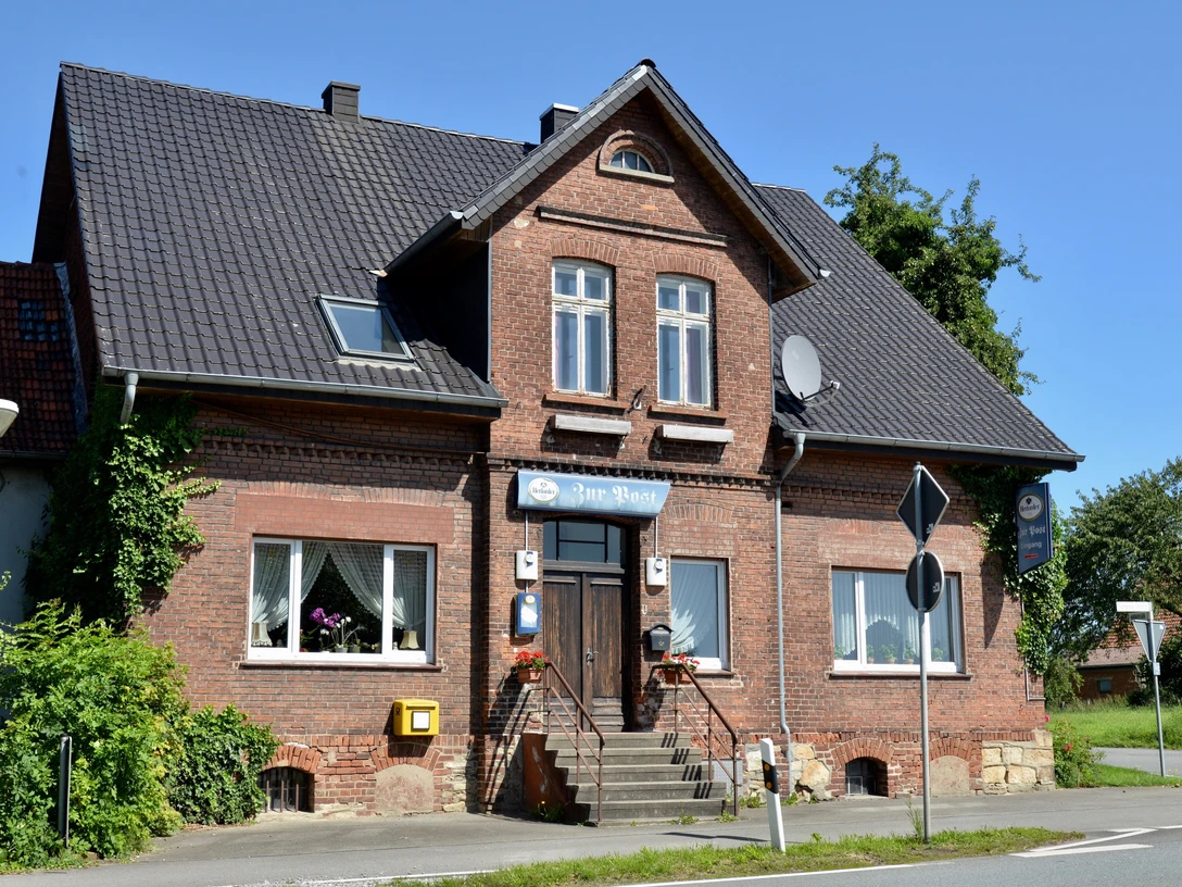 Restaurant Zur Post Backstein-Bau mit Ziegeldach und Treppe, zwei Fenster, umgeben von Pflanzen, unter blauem Himmel.