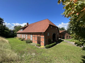Historisches Backsteinhaus mit rotem Satteldach, umgeben von grüner Wiese, unter blauem Himmel.