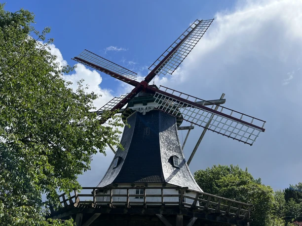 A windmill in the countryside with spinning blades under a blue sky, surrounded by trees and clouds.
