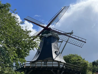 Schmomühle Mühle im Grünen mit funktionierenden Flügeln unter blauem Himmel, umgeben von Bäumen und Wolken.