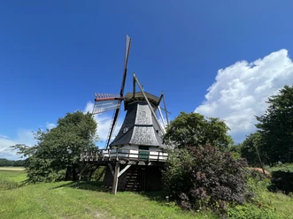 Schmomühle Historische Mühle mit Holzschindeldach und rot-weißen Flügeln, umgeben von Bäumen und blauem Himmel.