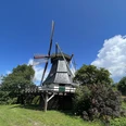 Schmomühle Historische Mühle mit Holzschindeldach und rot-weißen Flügeln, umgeben von Bäumen und blauem Himmel.