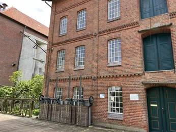 Backsteingebäude einer historischen Mühle mit großen Fenstern und Details, flankiert von grüner Vegetation.
