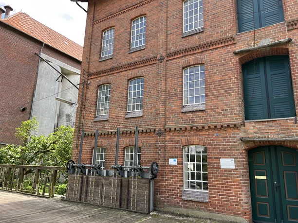 A brick building that is part of a historic mill, featuring large windows and intricate details, flanked by lush greenery.