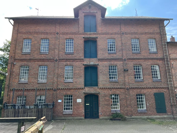 A brick watermill with green doors and windows, set in a flat, paved forecourt.