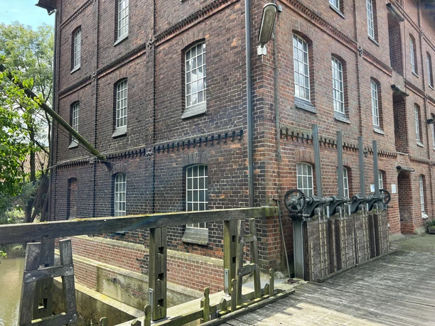 Alte Wassermühle mit roten Ziegeln und hölzernem Brückensteg, umgeben von üppigem Grün.