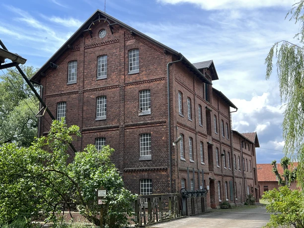 Wassermühle Stemmen Alte Wassermühle aus rotem Backstein mit fünf Etagen, umgeben von Bäumen und blauem Himmel.