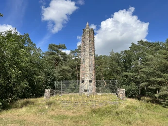 Lindhoopdenkmal Steinerner Obelisk in einer Waldlichtung, umgeben von Bäumen und unter einem blauen Himmel mit Wolken.