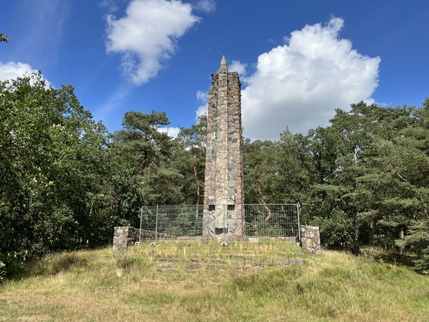 Lindhoopdenkmal Steinerner Obelisk in einer Waldlichtung, umgeben von Bäumen und unter einem blauen Himmel mit Wolken.
