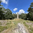 Langer Gedenkstein umgeben von Wiese und Bäumen unter strahlend blauem Himmel mit weißen Wolken.