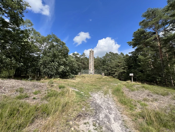 Langer Gedenkstein umgeben von Wiese und Bäumen unter strahlend blauem Himmel mit weißen Wolken.