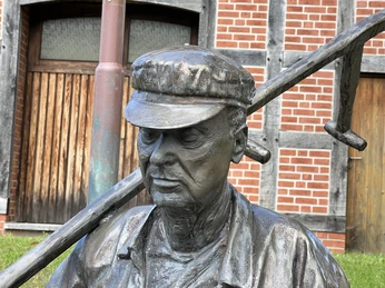 Bronzestatue eines älteren Mannes mit Schiebermütze, der einen Spaten über der Schulter trägt.