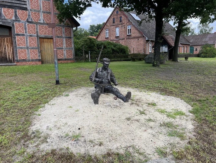 Skulptur „Scheepers Vadder“ Bronzestatue eines sitzenden Landarbeiters mit Axt, umgeben von historischen Fachwerkhäusern.