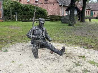 Bronzefigur eines sitzenden Mannes in Arbeitskleidung, der lächelnd eine Pfeife zwischen den Zähnen hält.