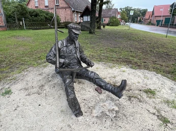 Eine Bronzefigur eines sitzenden Mannes beim Sägen auf einer Sandfläche neben einer Straße.