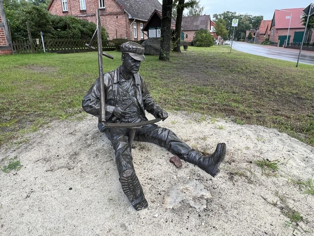Skulptur „Scheepers Vadder“ Eine Bronzefigur eines sitzenden Mannes beim Sägen auf einer Sandfläche neben einer Straße.