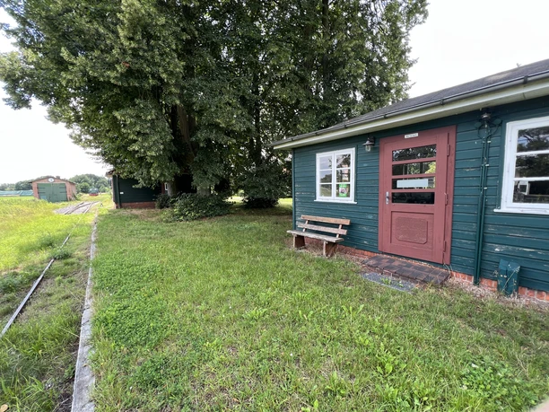 A green wooden building with a red door stands next to a meadow, surrounded by trees and a railroad line.
