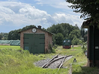An old goods shed with green gates and clock, next to it a rail track with a historic locomotive.