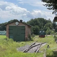 Ein alter Güterschuppen mit grünen Toren und Uhr, daneben ein Schienenweg mit einer historischen Lokomotive.An old goods shed with green gates and clock, next to it a rail track with a historic locomotive.En gammel godsbanegård med grønne porte og ur, ved siden af et jernbanespor med et historisk lokomotiv.Een oude goederenloods met groene hekken en klok, ernaast een treinspoor met een historische locomotief.