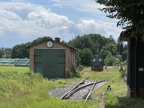 An old goods shed with green gates and clock, next to it a rail track with a historic locomotive.