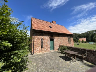 Rotes Ziegelhaus mit rotem Satteldach und grüner Tür, umgeben von einem gepflasterten Hof und Bäumen.