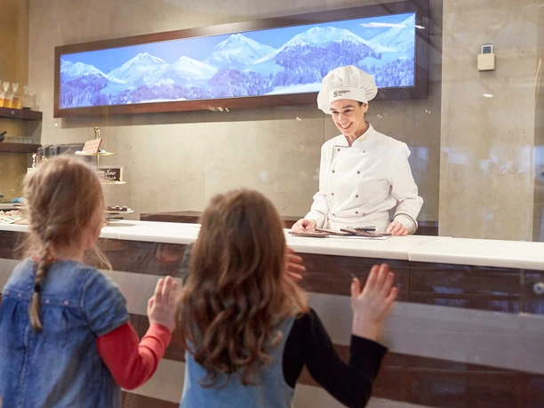 Schokoladenmuseum Eine Chocolatierin in weißer Uniform, umgeben von modernem Interieur, spricht mit drei Kindern.