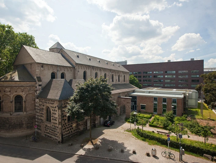 Museum Schnütgen Auf dem Bild ist das Museum Schnütgen in Köln zu sehen. Links erstreckt sich die romanische Cäcilienkirche, die Bruchsteinmauern und Rundbogenfenster zeigen. Rechts grenzt ein moderner Anbau an. Im Vordergrund ist ein kleiner Platz mit Bäumen und einigen Fahrrädern erkennbar.The picture shows the Schnütgen Museum in Cologne. On the left is the Romanesque Church of St. Cecilia, with its quarry stone walls and arched windows. On the right is a modern extension. A small square with trees and a few bicycles can be seen in the foreground.
